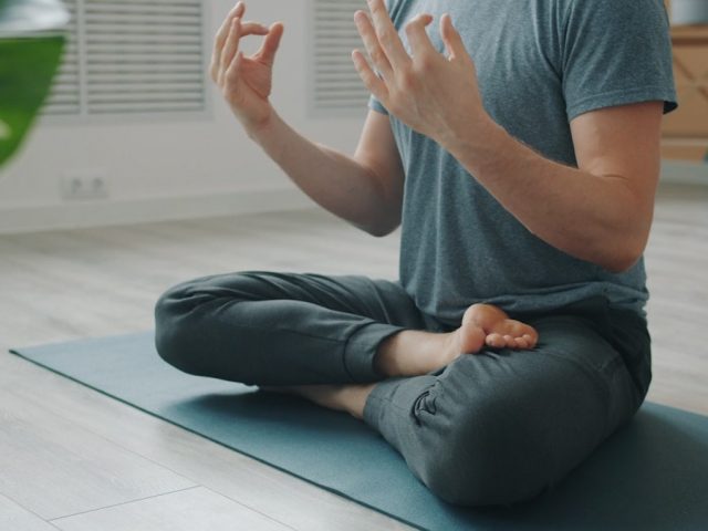 Low shot of anonymous man meditating on yoga mat sitting in lotus position with hands in namaste then in mudra gestures. People and relaxation concept.